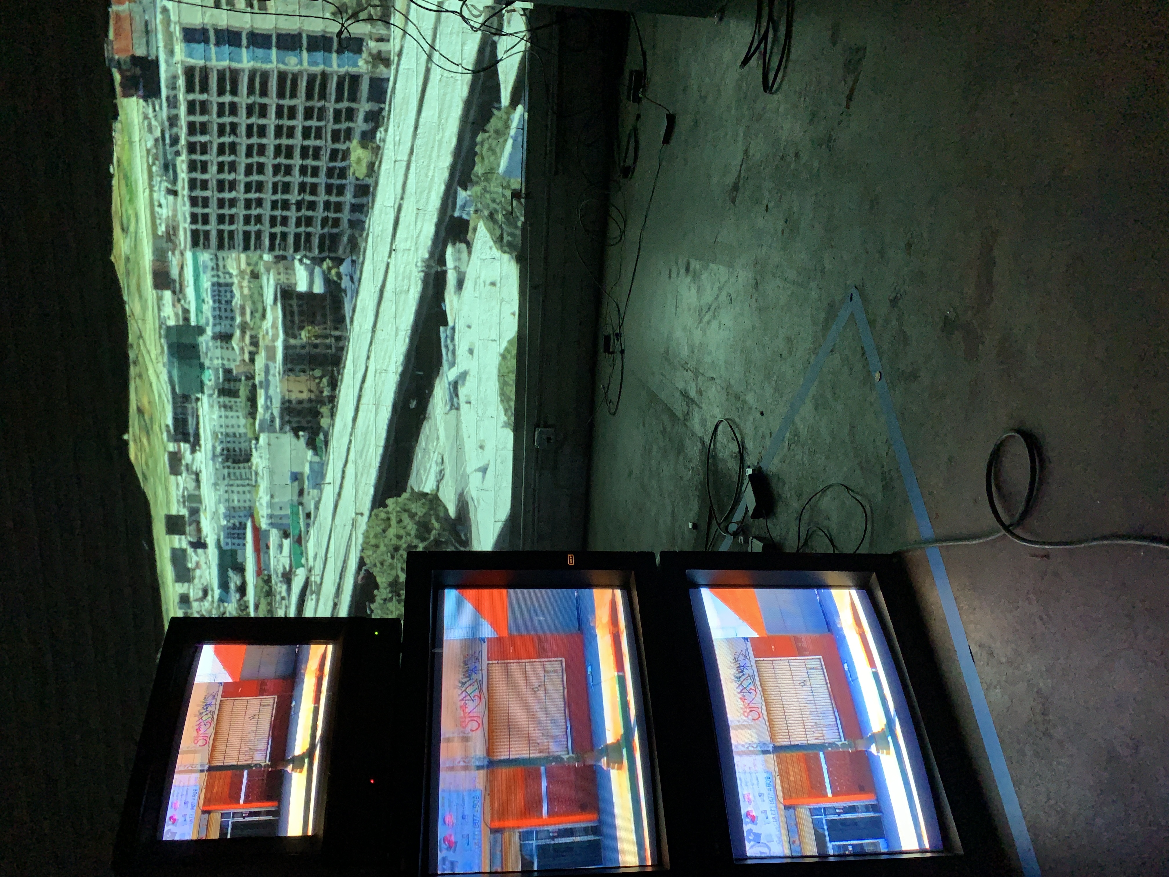 Stacked televisions in the foreground of the image depict an industrial corner in Los Angeles. In the background of the image is a projection of an aerial view of the city. They exist in a dark concrete and brick room.