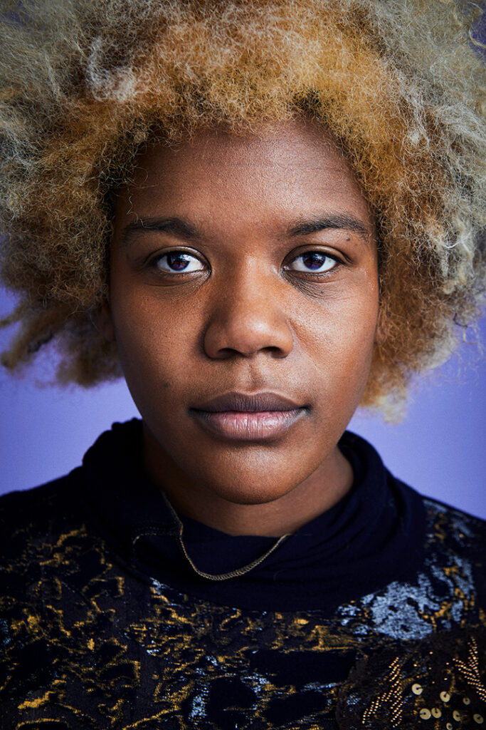 A photo of Samantha CC from the chest up. The artist casually gazes beyond the camera and is wearing a black mock neck sweater with gold designs. They are wearing a gold chain.