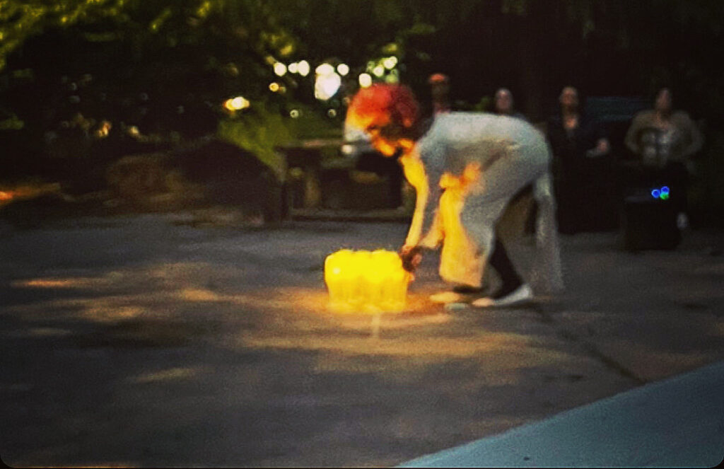 A blurry photo of the artist at mid distance. They are wearing a white robe, their hair is red. They are in a semi natural, semi concrete environment. They are bending over and reaching out their hands to a bright yellow glowing object on the ground. there is a small audience in the background.