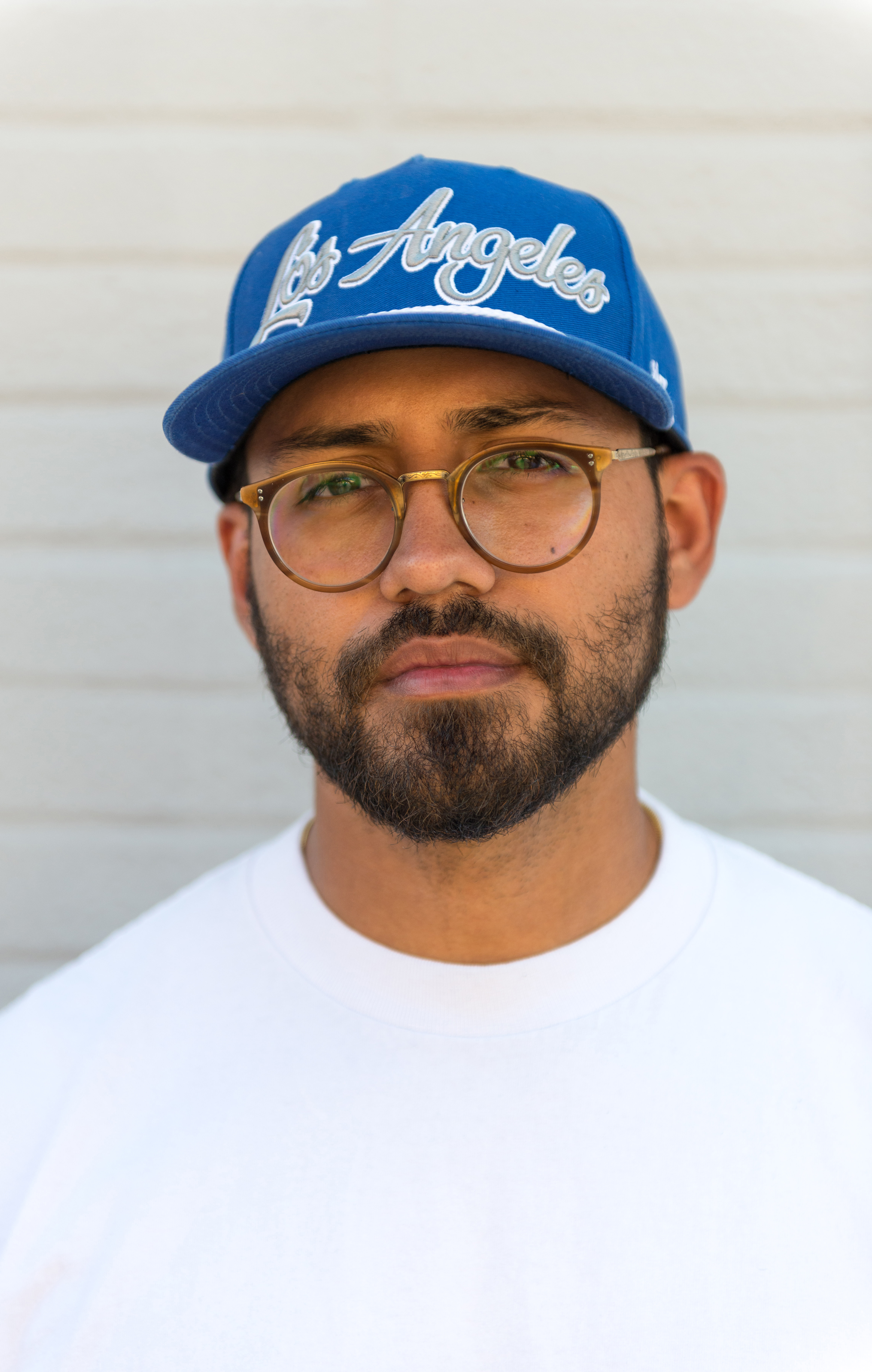 Shoulders-up photo of a person with tan/brown skin, a light beard and moustache, thin framed glasses and a blue hat that reads, "Los Angeles"