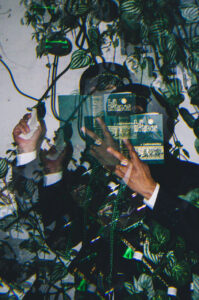 A double exposure of a person holding a book in front of their face as if proselytizing. They are in a suit with a naval hat and a ski mask on. There is foliage all around. 