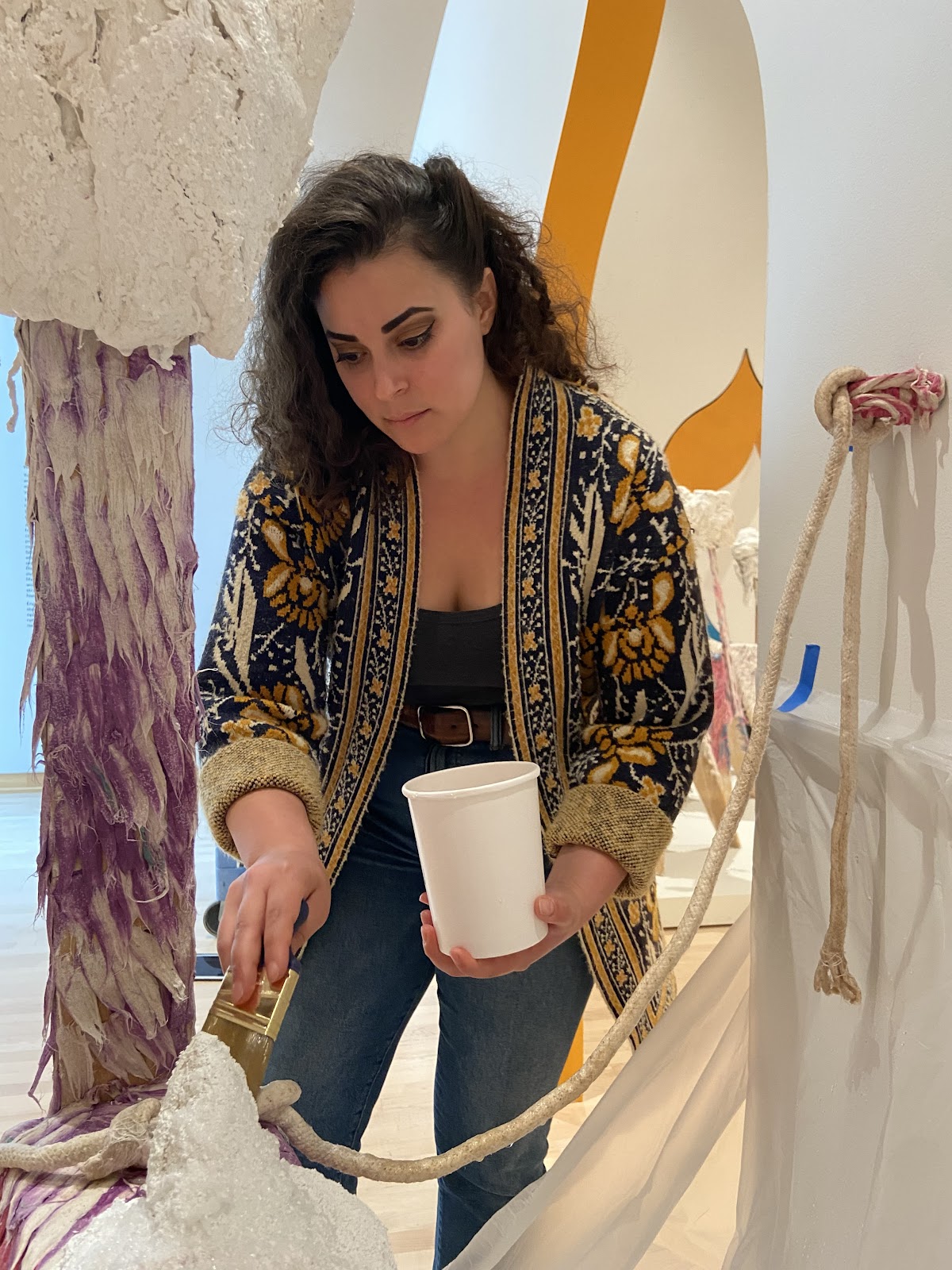 A photo of Yasmine Kasem holding a paint cup and a brush surrounded by sculpture and textile artworks.
