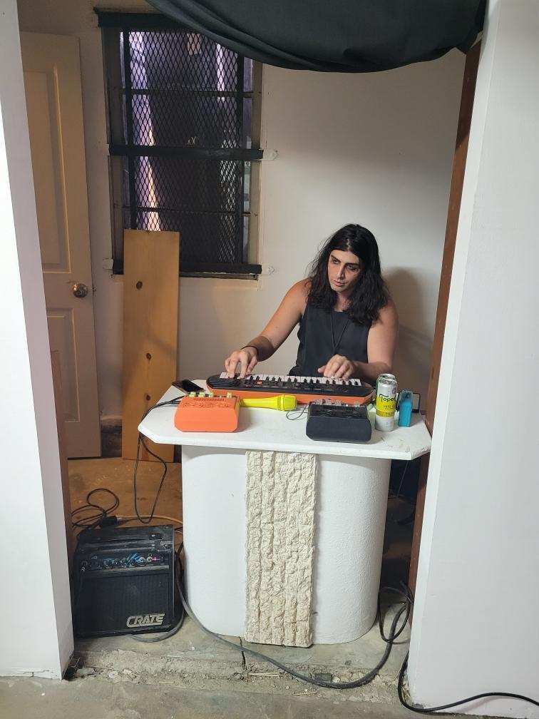 A person with long dark hair and sunken eyes sitting at a table playing a small keyboard