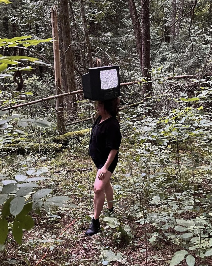 rachel jones, the artist, is standing in a daylight forest holding a television on her head which is displaying static